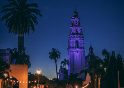 Giant palm tree and the architecture of the Balboa Park museums in the foreground with the colorfully lit California Tower in the background. Photo taken at dusk.